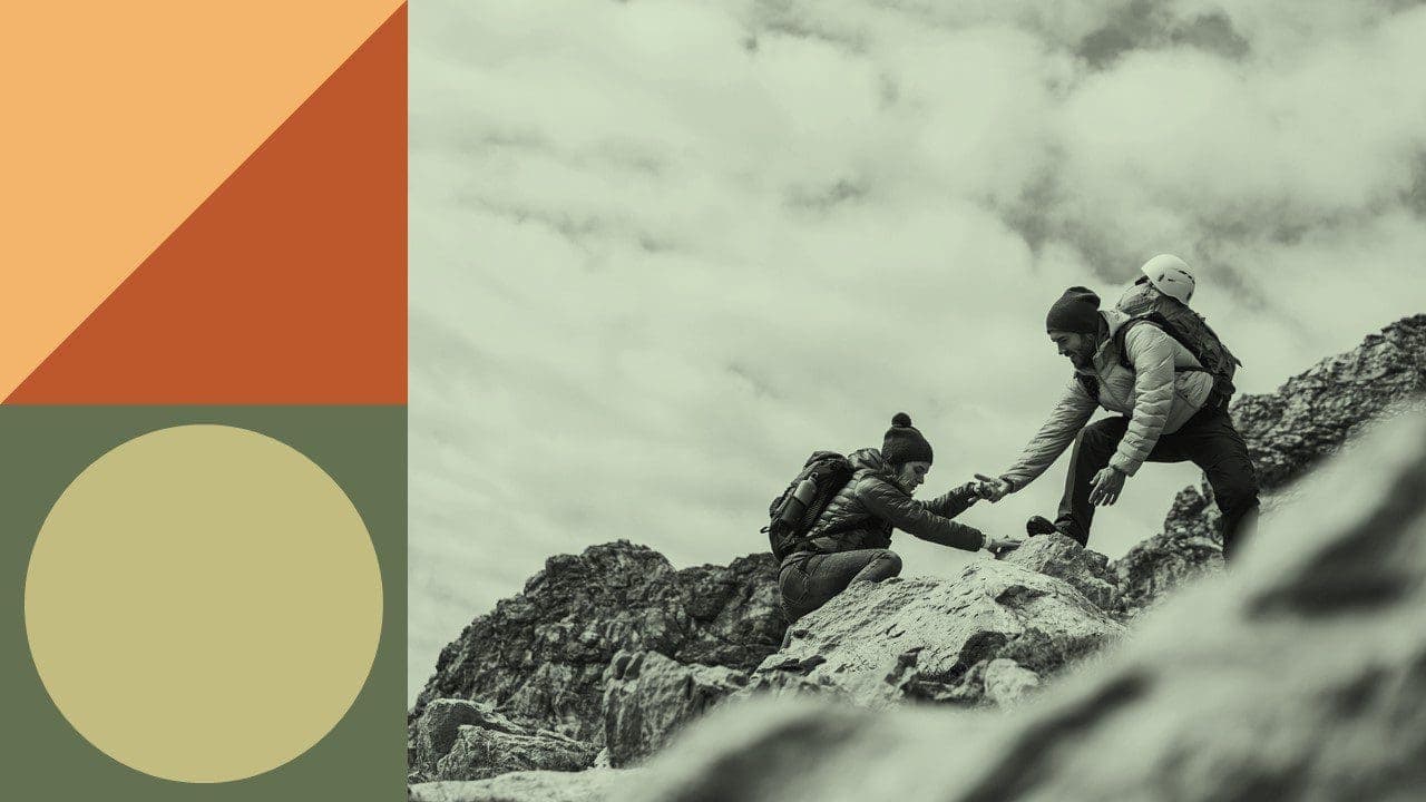 A hiker helping another hiker up a steep part of a mountain by holding their hand and leading.