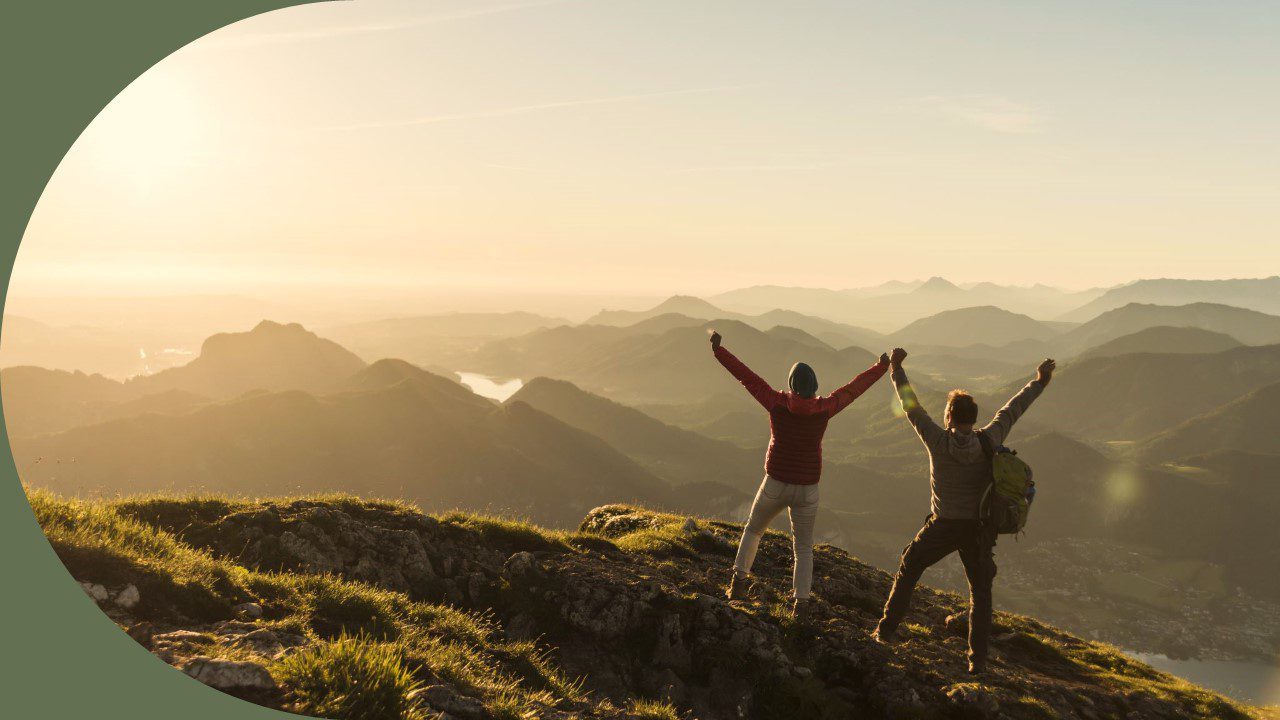 Two hikers on top of a mountain facing away from the camera and posing as by throwing their hands up in a cheering motion. They both look excited, relieved, and proud to have made the journey.